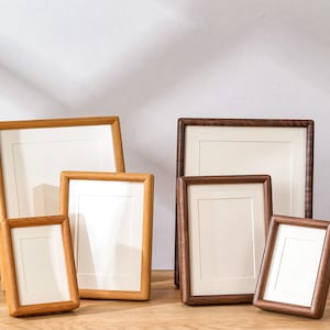 Puede incluir: Una colección de seis marcos de fotos rectangulares dispuestos en dos grupos. Tres marcos están hechos de madera clara y tres de madera oscura. Cada marco presenta un borde de cartón blanco que rodea un centro vacío.