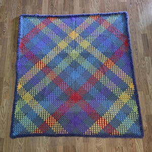 Könnte beinhalten: Eine rechteckige Häkeldecke mit einem mehrfarbigen Schottenmuster. Das Design besteht aus sich kreuzenden diagonalen Linien in den Farben Rot, Gelb, Grün, Blau und Violett, die einen Gittereffekt über die gesamte Oberfläche des Textils erzeugen.