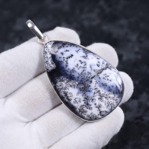 Könnte beinhalten: Ein tropfenförmiger Anhänger mit einem polierten dendritischen Opal, der eine weiße Basis und komplexe, farnartige schwarze Einschlüsse aufweist. Der Edelstein ist in eine glatte, silberfarbene Metallfassung mit einer passenden Öse gefasst.