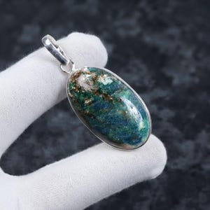 Puede incluir: Un colgante de forma ovalada que presenta una piedra preciosa pulida con inclusiones minerales en tonos azul oscuro, verde y marrón. La piedra está engastada en un bisel de metal plateado liso con una anilla plateada a juego en la parte superior.