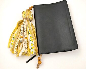 May include: A black book with a decorative tassel attached to the spine. The tassel features yellow and white ribbons, lemon-patterned fabric, and wooden beads with the text MY JESUS MAKING LEMONADE printed on square wooden blocks.