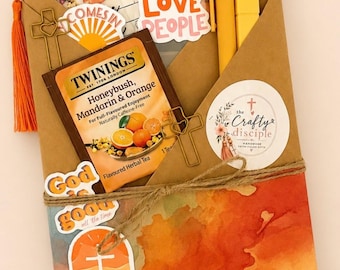 May include: A brown paper envelope containing a Twinings Honeybush, Mandarin & Orange tea bag, two yellow pens, and several stickers with text including Love God Love People, God is good, and set free. Two gold cross-shaped paper clips are attached.