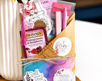 May include: A brown paper envelope filled with stationery items including a Twinings pomegranate and raspberry tea bag, a pink pen, a gold cross-shaped paperclip, and various floral stickers. The envelope is tied with twine and features colourful watercolour accents.