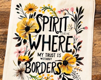 May include: A cream-coloured canvas tote bag featuring a floral design with yellow, pink, and black botanical illustrations. The text reads: SPIRIT LEAD ME WHERE MY TRUST IS WITHOUT BORDERS. The bag has two fabric handles at the top for carrying.