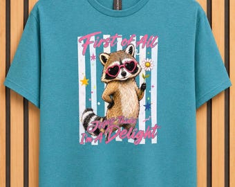 May include: A teal short-sleeved t-shirt featuring a graphic of a raccoon wearing pink heart-shaped sunglasses and holding a daisy. The text reads First of All Jesus Thinks I'm A Delight, surrounded by vertical white stripes and small colourful stars.