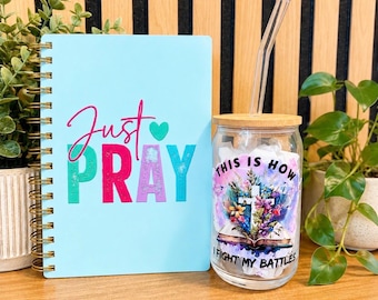 May include: A light blue spiral-bound notebook with the text Just PRAY and a small heart, alongside a clear glass tumbler with a bamboo lid, a glass straw, and a graphic of a cross, flowers, and the text THIS IS HOW I FIGHT MY BATTLES.
