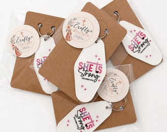 May include: A collection of white motel-style keychains featuring the pink and black text "SHE IS Strong" with "PROVERBS 31:25" and small star icons. Each keychain is attached to a brown card with a round "The Crafty Disciple" logo sticker.