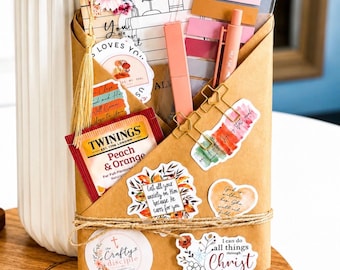May include: A brown paper envelope filled with stationery items including a Twinings Peach & Orange tea bag, a gold tassel bookmark, pens, sticky notes, and various stickers with religious text and floral designs, all secured with twine.