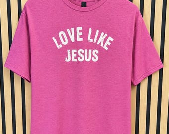 May include: A heathered pink short-sleeve t-shirt featuring the white distressed text LOVE LIKE JESUS printed in an arched design across the chest. The garment has a crew neckline and is displayed on a wooden hanger against a slatted wall.