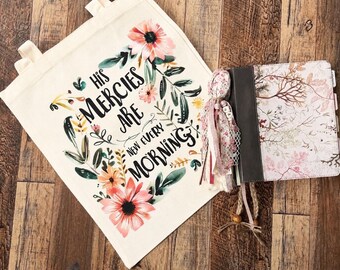 May include: A cream-coloured canvas tote bag featuring a floral wreath design and the black text "HIS MERCIES ARE NEW EVERY MORNING" alongside a square journal with a floral cover, a dark spine, and decorative fabric tassels attached to the side.