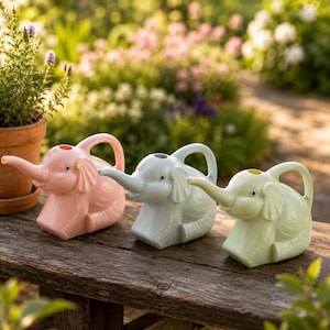 Pode incluir: Três regadores em forma de elefante nas cores rosa pastel, azul claro e verde pálido. Cada regador apresenta um bico longo em forma de tromba, uma alça na parte traseira e olhos móveis. Os itens estão alinhados sobre uma superfície de madeira.