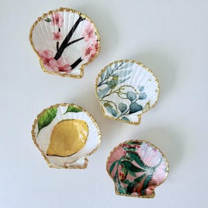 May include: Four decorative scallop shells with gold-painted edges. One shell features pink cherry blossoms on black branches, one has blue-green eucalyptus leaves, one displays a yellow lemon with a green leaf, and one shows pink floral patterns.