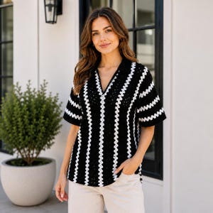 Könnte beinhalten: Ein kurzärmeliges Häkel-Poloshirt mit vertikalem Streifenmuster in Schwarz und Weiß. Das Kleidungsstück hat einen V-Ausschnitt mit Kragen und eine strukturierte, grob gestrickte Optik. Das Oberteil wird mit einer hellen Hose kombiniert.