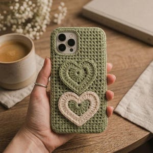 Könnte beinhalten: Eine salbeigrüne Handyhülle aus Häkelarbeit mit einem strukturierten Strickmuster und zwei herzförmigen Applikationen. Das obere Herz ist einfarbig salbeigrün, während das untere Herz eine salbeigrüne Mitte mit einem cremefarbenen Rand hat.