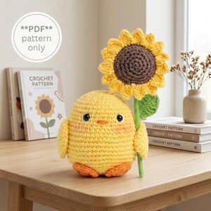 Könnte beinhalten: Ein gelbes gehäkeltes Küken-Stofftier, das eine große gelbe und braune Sonnenblume hält. Das Küken hat schwarze Augen, einen orangefarbenen Schnabel und orangefarbene Füße. Ein weißes Etikett zeigt **PDF** pattern only, Bücher im Hintergrund.