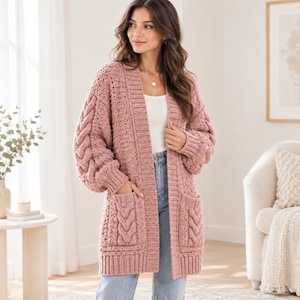Könnte beinhalten: Eine altrosa Strickjacke mit offener Vorderseite, grober Strickstruktur, Zopfmusterdetails an den Ärmeln und Vordertaschen, gerippten Abschlüssen an Kragen und Saum sowie langen Ärmeln. Das Kleidungsstück wird über einem weißen Oberteil und Jeans getragen.