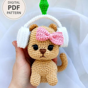 Könnte beinhalten: Eine beige Häkelkatzenpuppe mit einer rosa Schleife, weißen Kopfhörern und einem grünen rautenförmigen Aufsatz. Die Katze hat schwarze Augen und eine braune Nase. Ein kreisförmiges Abzeichen oben links zeigt den Text DIGITAL PDF PATTERN.