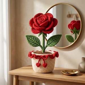 Puede incluir: Una rosa de ganchillo hecha a mano con pétalos de color rojo vibrante y un tallo verde con dos hojas. La flor está plantada en un recipiente de ganchillo beige con un borde rojo con volantes, un lazo decorativo rojo y dos corazones.