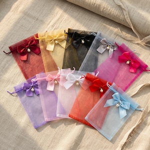 Könnte beinhalten: Eine Sammlung von zehn rechteckigen Organza-Geschenkbeuteln mit Kordelzug in verschiedenen Farben wie Rot, Gold, Schwarz, Silber, Pink, Lila, Hellrosa, Weiß, Orange und Blau. Jeder Beutel hat eine passende Satin-Schleife mit einer Perle.