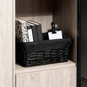 May include: A rectangular black woven wicker basket with a black fabric liner. Inside the basket are three books, one with a black and white floral pattern and two with solid black covers, alongside a black metal clip holding a white note card.