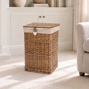May include: A rectangular brown wicker laundry hamper with a matching lid. The hamper features a beige fabric liner that folds over the top edge and a brown leather strap with a small white heart-shaped wooden tag attached to the front.