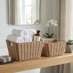 May include: Two rectangular woven seagrass baskets with integrated handles. The larger basket contains three rolled white towels and a brown glass bottle of bath oil with a white label. A small bar of soap and a succulent plant are nearby.