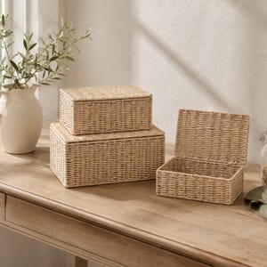 May include: Three rectangular storage boxes crafted from light beige woven material. Two boxes are stacked on top of each other, while the third box sits to the right with its hinged lid open, revealing the hollow interior of the woven container.
