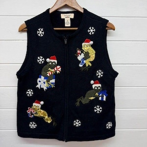 May include: A black knit zip-up vest featuring embroidered green frogs wearing red and white Santa hats. The frogs are depicted holding candy canes and gift boxes, surrounded by white embroidered snowflakes. The vest has a label reading bechamel.