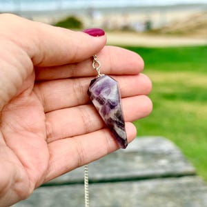 May include: A faceted, pointed amethyst crystal pendant with deep purple and white marbled patterns. The stone is attached to a small silver-toned metal bail and hangs from a thin silver-toned chain, displaying a polished, smooth surface texture.