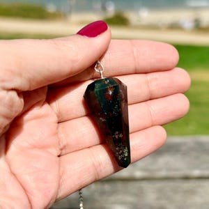 May include: A faceted, pointed pendulum made of dark green bloodstone with visible red and white mineral inclusions. The stone is suspended from a thin silver-colored metal chain, featuring a polished surface with a geometric, multi-sided shape.