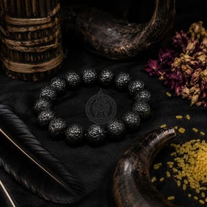 Könnte beinhalten: Ein kreisförmiges Armband aus dunklen, texturierten, kugelförmigen Perlen mit einer porösen, vulkanartigen Oberfläche. Die Perlen sind zu einer durchgehenden Schlaufe aufgereiht und zeigen ein schwaches, kreisförmiges geometrisches Emblem.