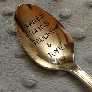 Puede incluir: Una cuchara de plata con las palabras "PLANES TRAINS TRUCKS & TOYS" grabadas en la parte delantera.
