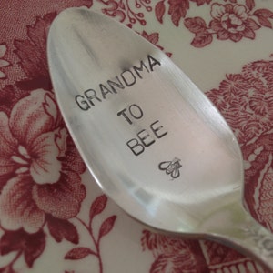 May include: A silver spoon with the words "GRANDMA TO BEE" etched on the bowl, along with a small bee illustration. The spoon rests on a red and white floral patterned surface.