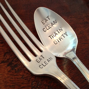 Puede incluir: Un tenedor y una cuchara de plata con las palabras "Eat Clean Train Dirty" grabadas en ellos. El tenedor tiene las palabras "Eat Clean" grabadas en él, y la cuchara tiene las palabras "Train Dirty" grabadas en ella.