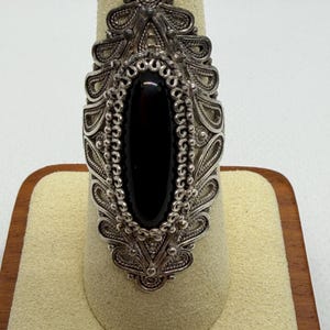 May include: A silver ring featuring an elongated oval black stone set in the center. The metalwork surrounding the stone displays intricate filigree patterns with small silver beads, creating a detailed, ornate design that extends vertically.