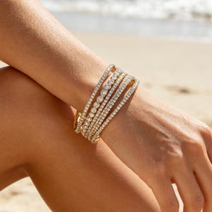May include: A set of five gold-toned tennis bracelets worn together on a wrist. Each bracelet features a continuous row of clear, round-cut stones set in a metallic band, creating a layered, sparkling appearance across the forearm.