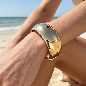 May include: A wide, polished gold-tone cuff bracelet with a smooth, curved surface. The metal has a high-shine finish that reflects the surrounding environment, creating a bright, metallic appearance on the wrist of the wearer.