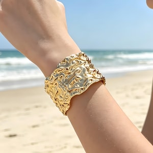 Può includere: Un ampio bracciale a fascia color oro caratterizzato da una superficie altamente strutturata, irregolare e stropicciata che riflette la luce. Il cinturino in metallo ha una finitura lucida con profonde rientranze e creste in rilievo.