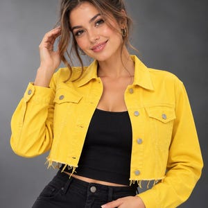 May include: A bright yellow cropped denim jacket featuring a classic collar, long sleeves with buttoned cuffs, two chest pockets with button flaps, and a front button closure. The hem of the jacket is finished with a raw, frayed edge detail.