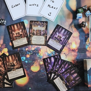 Könnte beinhalten: Ein Tischspiel-Setup mit mehreren Spielkarten, darunter Karten mit der Aufschrift Emperor und General, sowie verstreuten mehrseitigen Würfeln. Drei weiße Karten oben zeigen den Text Enjoy Ways To Play mit einem Smiley.