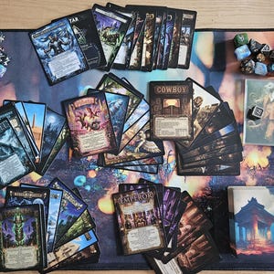 Può includere: Un set da gioco da tavolo con numerose carte illustrate, dadi poliedrici multicolori e un grande tappetino rettangolare con un design astratto. Tre carte bianche riportano il testo Enjoy New Ways To Play! scritto in nero.