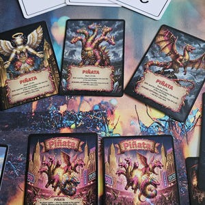 Pode incluir: Uma coleção de cartas de jogo retangulares com ilustrações de fantasia de dragões de várias cabeças e piñatas. Três cartas brancas no topo exibem o texto manuscrito "ENJOY", "WAYS" e "PLAY!" com um ícone de carinha feliz.