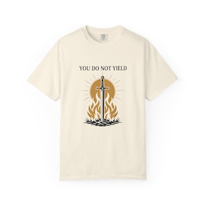 May include: A cream-colored short-sleeve t-shirt featuring a graphic print of a sword standing upright in a mound of earth, surrounded by stylized flames and a sunburst. The text "YOU DO NOT YIELD" is printed in black above the sword illustration.