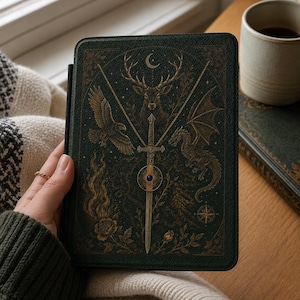 May include: A dark green e-reader case featuring a gold-toned illustration of a central sword with a blue gemstone, flanked by a flying eagle, a stag, and a dragon. The design includes a crescent moon, floral elements, and a compass rose.