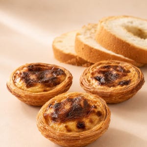 May include: Three round egg tarts with golden-brown, flaky pastry crusts and caramelized, blistered custard centers, accompanied by two slices of round, light-colored bread positioned in the background on a neutral-toned surface.