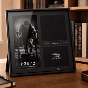 Puede incluir: Un marco de exhibición negro que contiene una fotografía en blanco y negro de un atleta empujando un trineo, el texto 1:34:12 OFFICIAL FINISH TIME, una placa con el nombre SIMON y un emblema metálico HYROX sobre un fondo de tela negra.