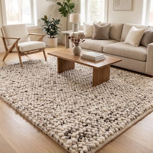 May include: A rectangular area rug featuring a thick, chunky knit texture made of cream-colored yarn with interspersed flecks of dark gray and black fibers, creating a dense, nubby surface that covers the floor in a living room setting.