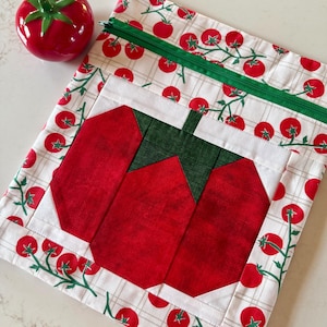 May include: A fabric pouch featuring a white background with a red tomato and green vine print, a green zipper, and a large central red tomato quilt block design. Beside the pouch sits a small, round, red ceramic tomato-shaped object.