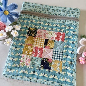 May include: A rectangular fabric zipper pouch featuring a central patchwork square made of various floral, gingham, and solid-colored fabric blocks. The surrounding fabric is a light blue print with a repeating pattern of small bees and flowers.