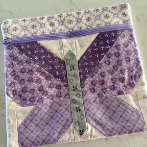 May include: A rectangular fabric pouch featuring a quilted butterfly design in shades of purple, lavender, and cream. The pouch includes a top zipper closure in a matching lavender hue and a central gray body segment with a small geometric pattern.